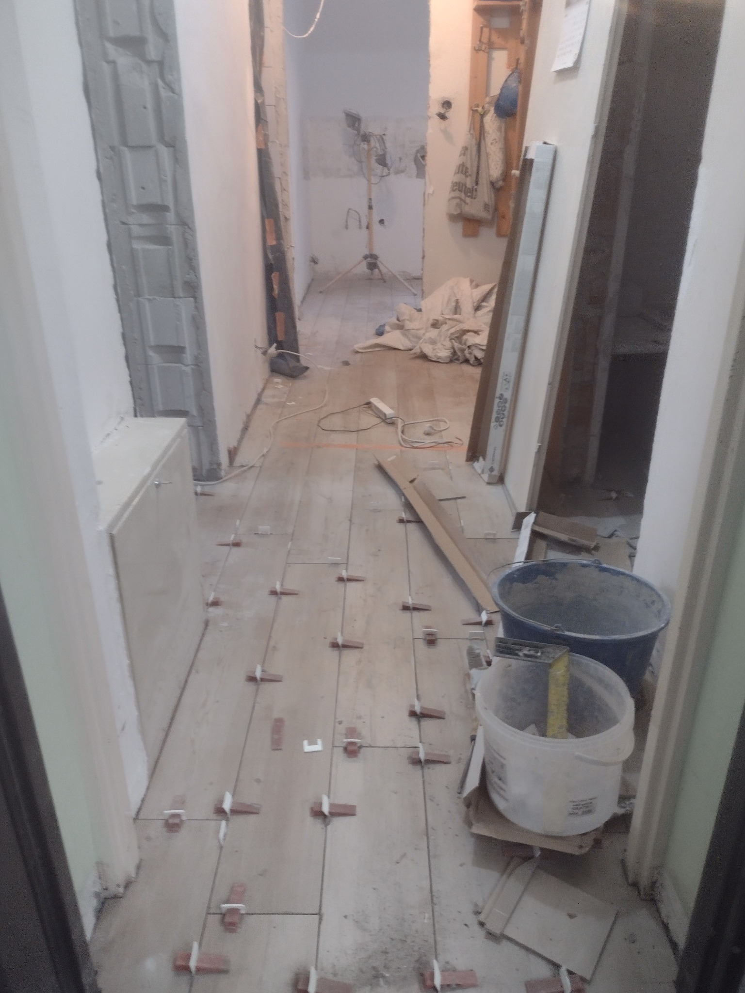 Układanie płytek podłogowych w korytarzu, widoczne krzyżyki dystansowe, dwa wiadra budowlane, narzędzia i materiały budowlane w tle.