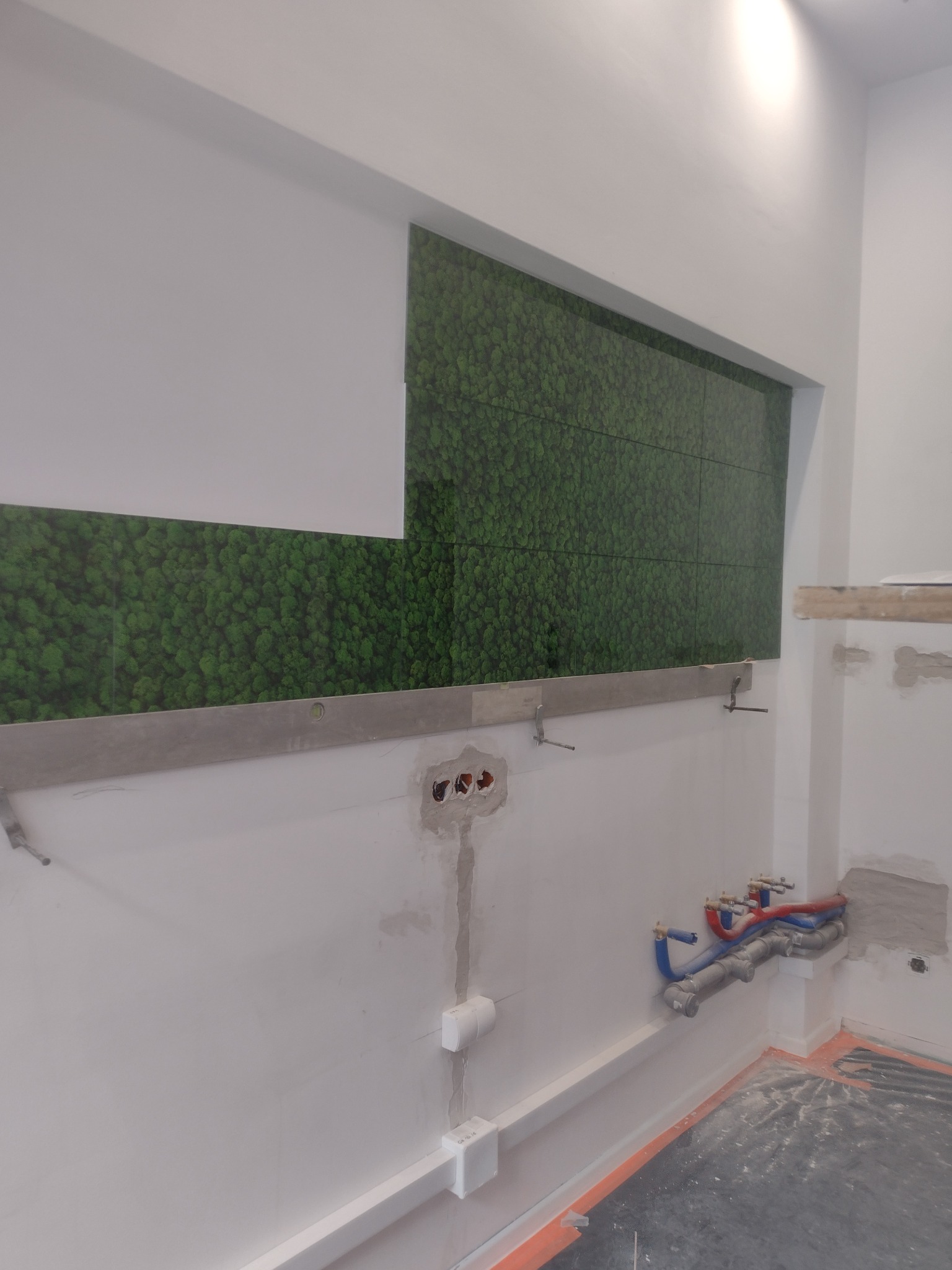Ściana łazienki w trakcie prac wykończeniowych z fragmentem dekoracyjnego panelu imitującego mech, widoczne instalacje wodne i elektryczne oraz poziomica z uchwytami montażowymi.