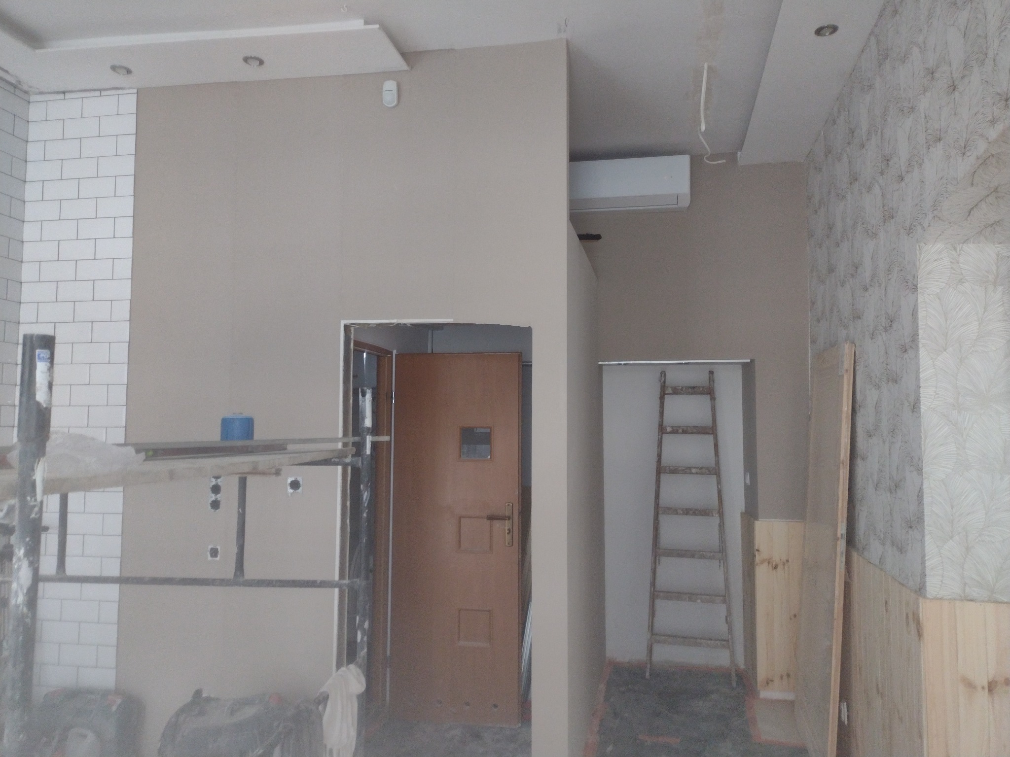 Wnętrze w trakcie prac wykończeniowych: fragment ściany z białymi płytkami, pomalowana na beżowo ściana z otworami na gniazdka elektryczne, drzwi wewnętrzne, drabina oparta o ścianę pokrytą tapetą...