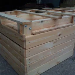 Nowe, jasne skrzynio-palety z surowego drewna, skręcone śrubami, ułożone jedna na drugiej na betonowym podłożu.