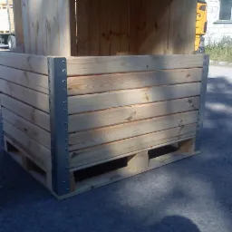 Skrzyniopaleta z jasnego drewna, wzmocniona metalowymi narożnikami, ustawiona na asfalcie; w tle widoczny samochód dostawczy i budynek.