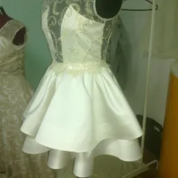 Biała, krótka suknia z tiulową górą, zdobiona haftem i koralikami, prezentowana na manekinie krawieckim w pracowni.