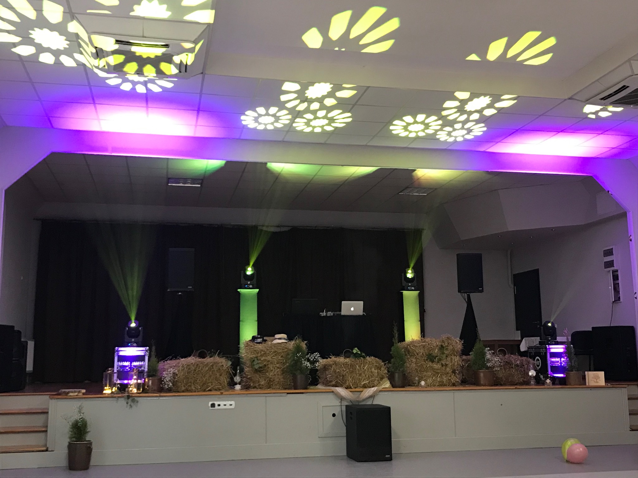 Scena udekorowana na imprezę z belami siana, roślinami w doniczkach, reflektorami z zielonymi promieniami, sprzętem DJ-skim i dekoracyjnym oświetleniem sufitowym w kształcie kwiatów.