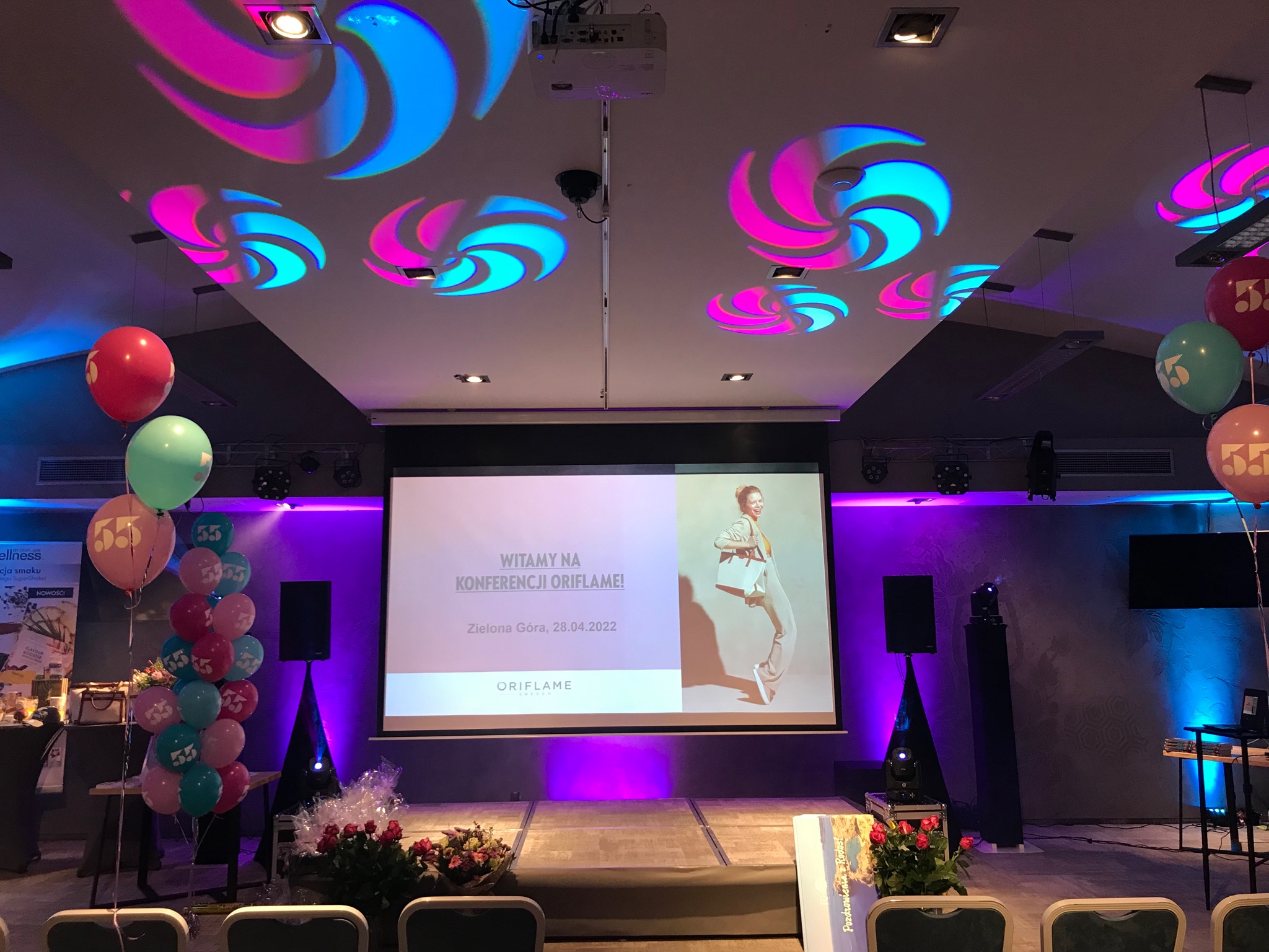 Sala konferencyjna z projekcją na ekranie 'Witamy na konferencji Oriflame!', dekoracje balonowe z numerem 55, reflektory na suficie tworzące wzory w kolorach różu i błękitu, nagłośnienie po bokach...