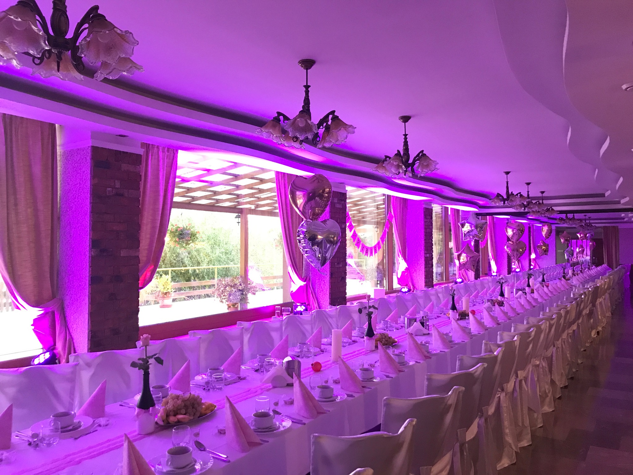 Uroczysta sala weselna w Międzychodzie, oświetlona fioletowymi lampami, z długim stołem nakrytym białymi obrusami i różowymi serwetkami, dekoracje z balonów w kształcie serc, widoczne żyrandole.