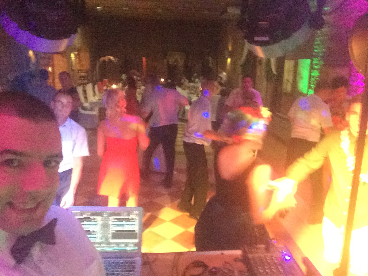 DJ w muszce uśmiecha się do kamery, w tle tańczący goście weselni na parkiecie oświetlonym kolorowymi światłami dyskotekowymi.