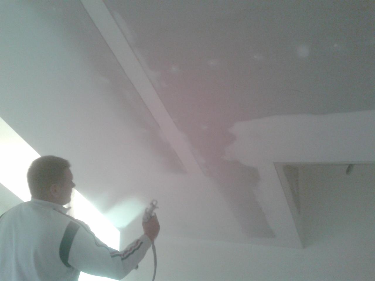 Pracownik maluje sufit natryskowo, widoczne niedomalowane fragmenty i narożnik pomieszczenia.