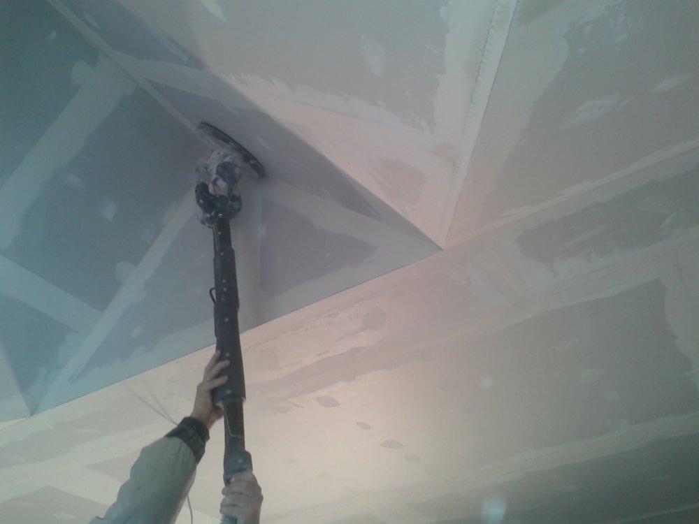 Szlifowanie gładzi gipsowej na suficie przy użyciu szlifierki teleskopowej, widoczne łączenia płyt gipsowo-kartonowych.