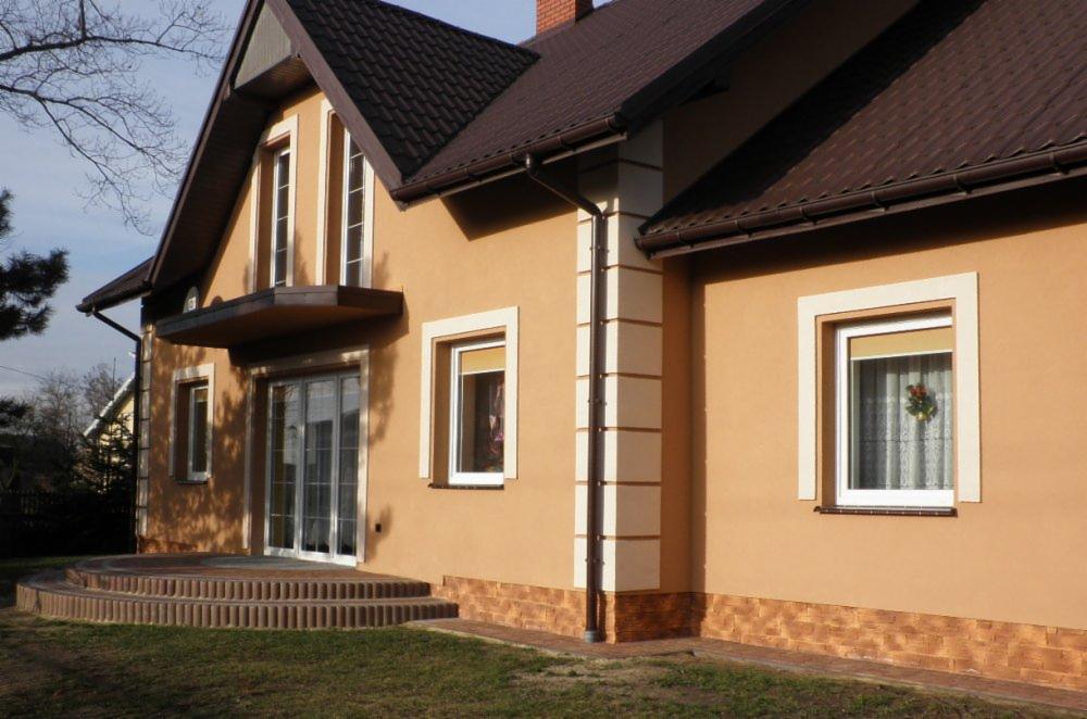 Ocieplony dom jednorodzinny z brązową dachówką, widoczne detale wykończenia elewacji, okna z dekoracją świąteczną, oraz schody wejściowe z kostki.