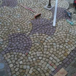 Układanie wzoru z kostki granitowej w odcieniach szarości i fioletu, widoczne narzędzia: miotła, poziomica i ubijak, teren w trakcie zagospodarowania.