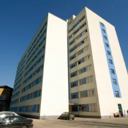 Wysoki budynek z licznymi oknami z niebieskimi ramami, widziany z dołu, z samochodami i zielenią w tle. Jasne, słoneczne oświetlenie.