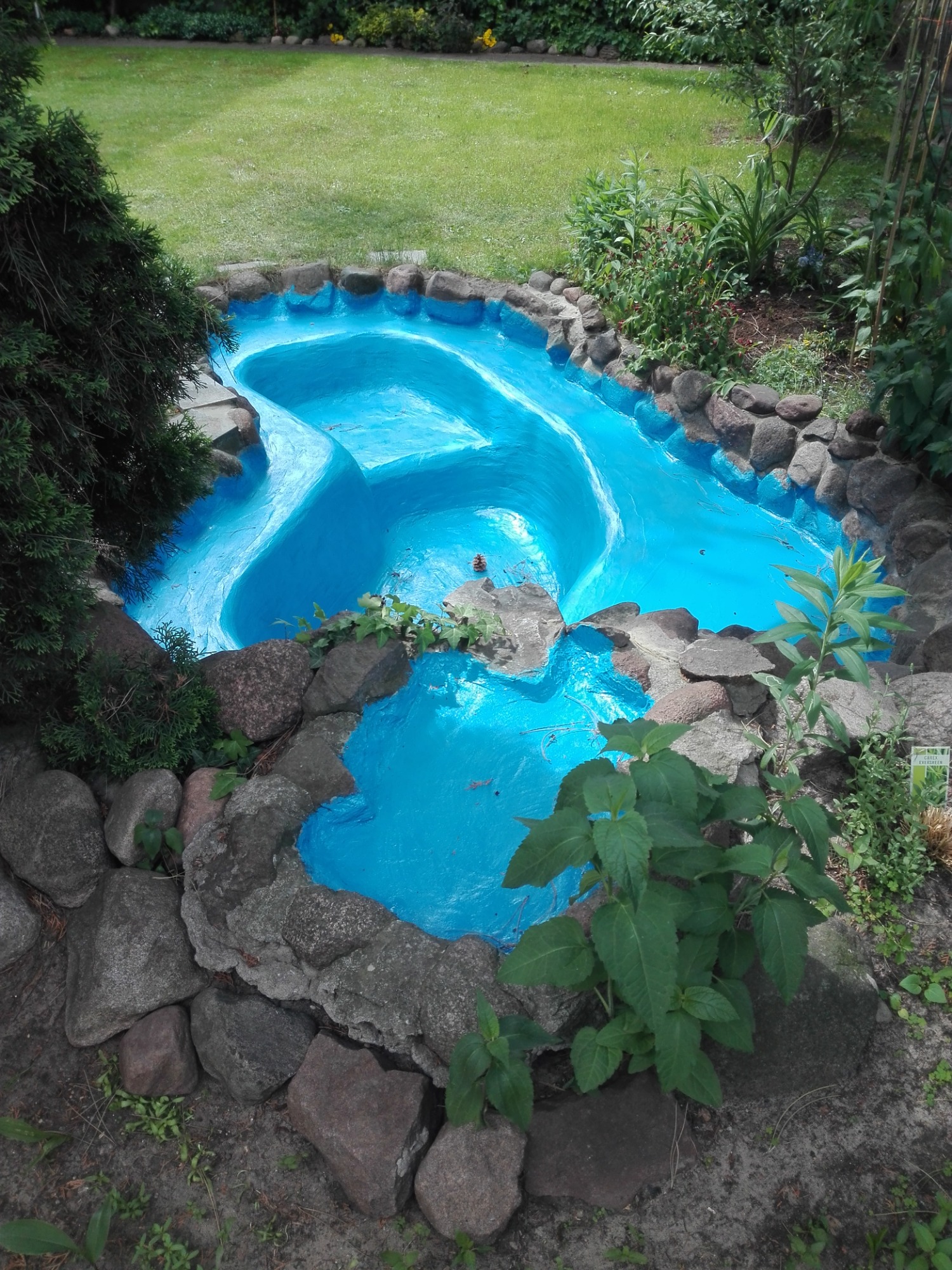 Ogród z małym, sztucznym jeziorkiem o nieregularnym kształcie, wyłożonym kamieniami i pomalowanym na intensywny, błękitny kolor, otoczonym zielenią trawnika i roślinności.