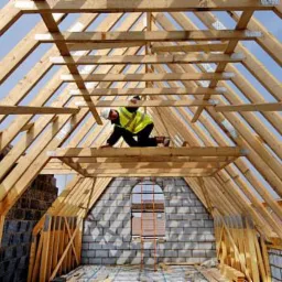 Pracownik w odblaskowej kamizelce montuje drewnianą konstrukcję dachu w surowym stanie, widok z wnętrza budynku na niebieskie niebo przez szkielet dachu.