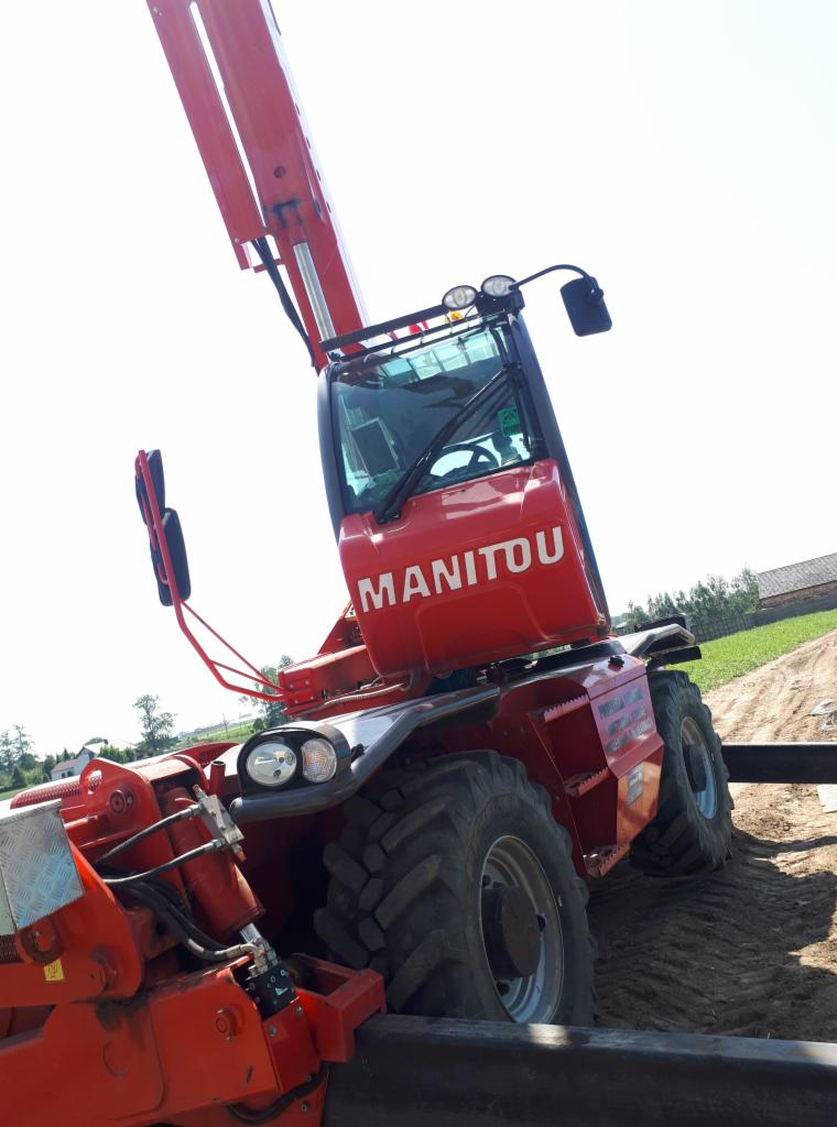 Czerwony podnośnik teleskopowy Manitou na placu budowy, częściowo widoczne elementy konstrukcji, zieleń w tle, pochylony kadr.