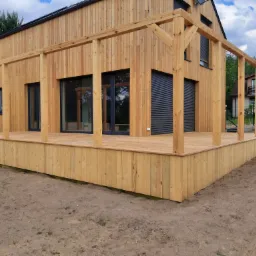 Nowoczesny dom z jasnego drewna z dużym tarasem i drewnianą pergolą w surowym stanie, z widocznymi czarnymi roletami zewnętrznymi.