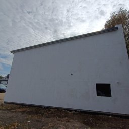 Lootus - Szara, otynkowana ściana budynku z małym, kwadratowym oknem, widoczna z niskiej perspektywy na tle pochmurnego nieba i jesiennych liści.