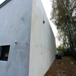 Lootus - Surowa elewacja budynku w trakcie wykańczania, z widocznymi przewodami elektrycznymi, oknem z czarną ramą i fragmentem drzewa z opadającymi liśćmi.