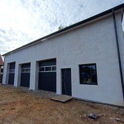 Lootus - Nowo otynkowany budynek gospodarczy z trzema bramami garażowymi w kolorze antracytowym, drzwiami wejściowymi i oknem, teren wokół jeszcze niezagospodarowany.