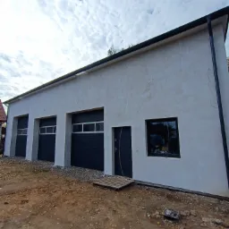 Nowo otynkowany budynek gospodarczy z trzema bramami garażowymi w kolorze antracytowym, drzwiami wejściowymi i oknem, teren wokół jeszcze niezagospodarowany.