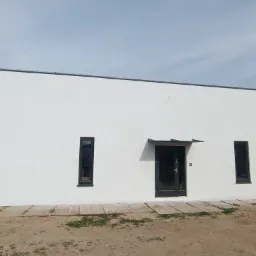 Biała elewacja budynku z ciemnymi oknami i drzwiami. Widoczne deski i piasek u podstawy, sugerujące prace wykończeniowe. Minimalistyczny design.