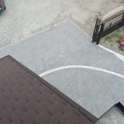 Widok z góry na szarą kostkę brukową z białymi wstawkami układającymi się w łuk, brązowy dach z bitumicznej dachówki, szary samochód i plac zabaw dla dzieci