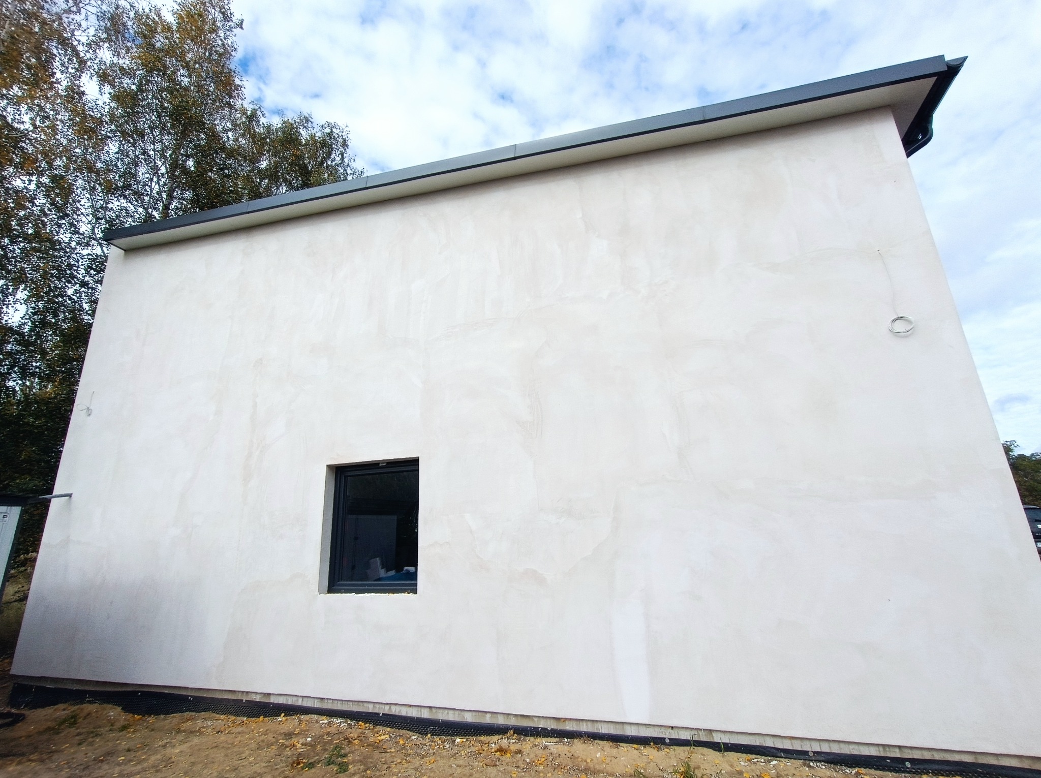 Nowoczesny, otynkowany budynek z małym, kwadratowym oknem i ciemnym dachem, widok z zewnątrz na tle nieba i drzew.