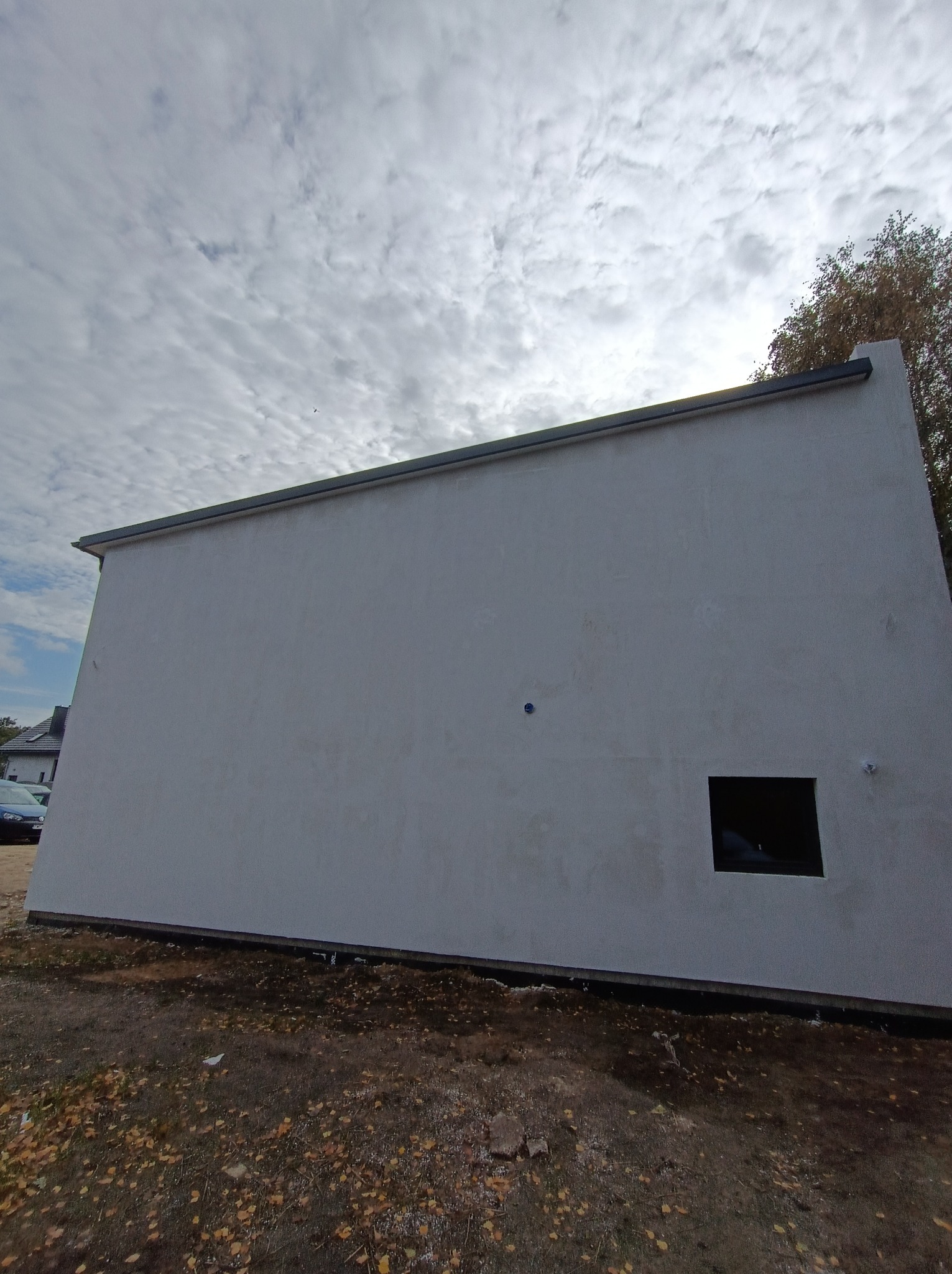 Szara, otynkowana ściana budynku z małym, kwadratowym oknem, widoczna z niskiej perspektywy na tle pochmurnego nieba i jesiennych liści.