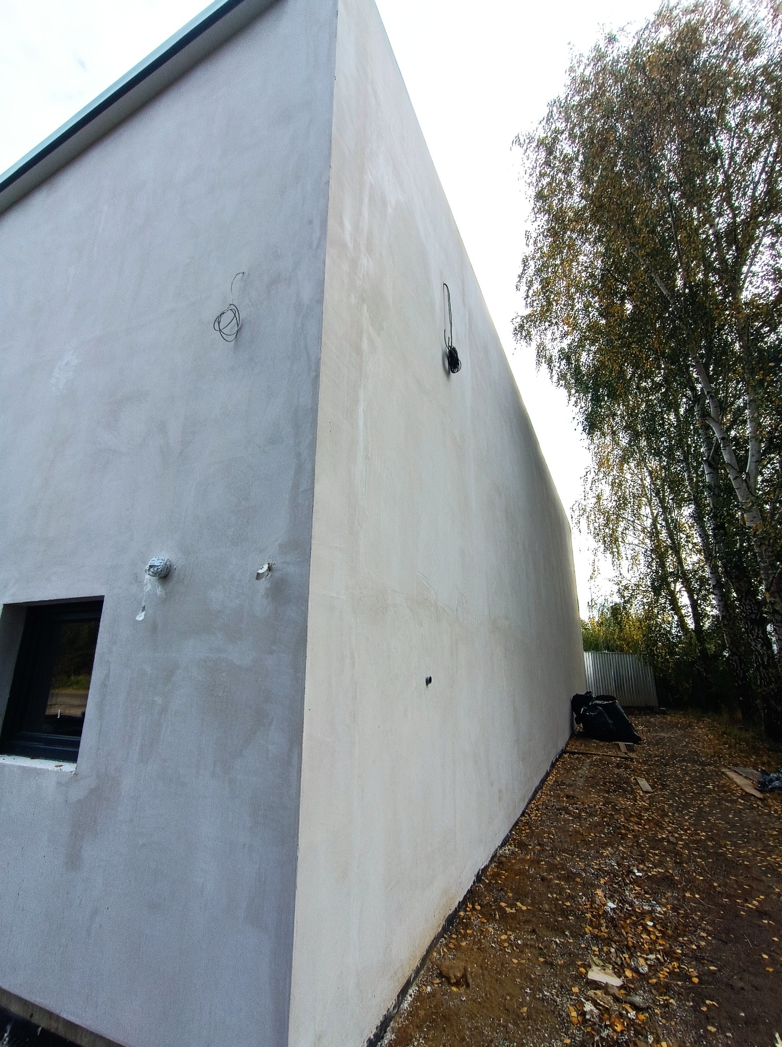Surowa elewacja budynku w trakcie wykańczania, z widocznymi przewodami elektrycznymi, oknem z czarną ramą i fragmentem drzewa z opadającymi liśćmi.