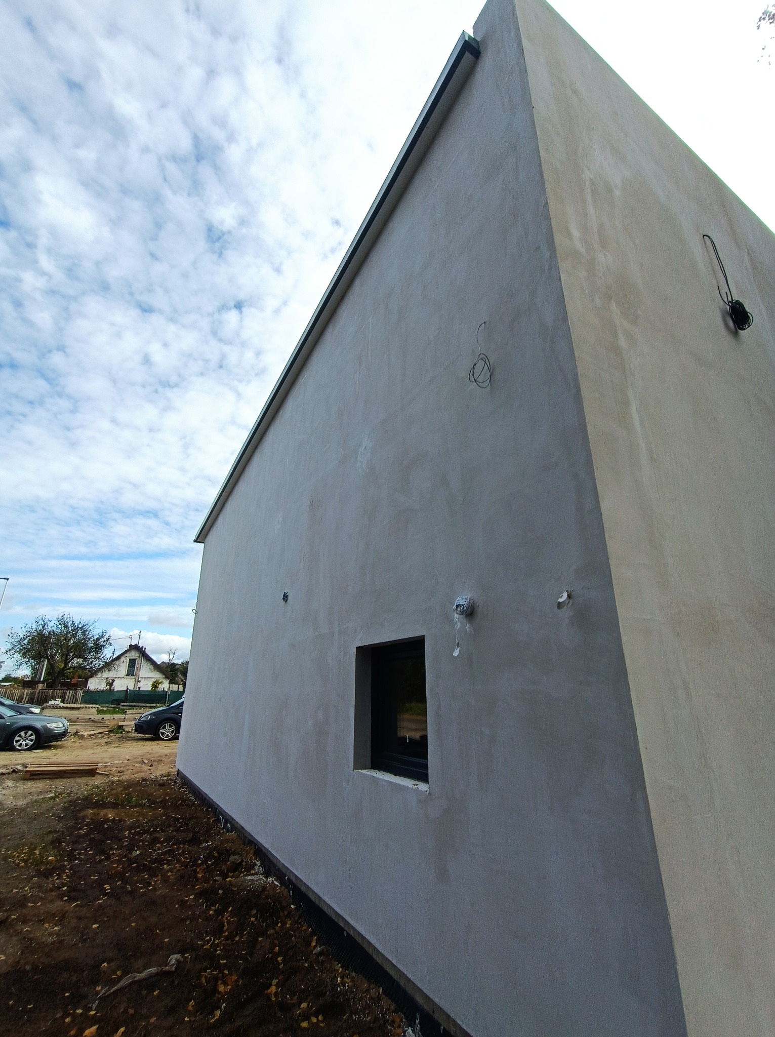 Świeżo otynkowana ściana budynku jednorodzinnego z czarną stolarką okienną, widoczne wystające przewody elektryczne, zarysowana linia podziału elewacji, w tle zabudowa mieszkalna i pochmurne niebo.