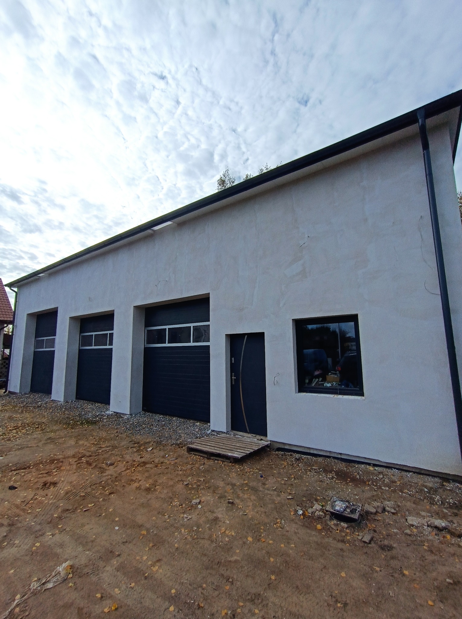 Nowo otynkowany budynek gospodarczy z trzema bramami garażowymi w kolorze antracytowym, drzwiami wejściowymi i oknem, teren wokół jeszcze niezagospodarowany.