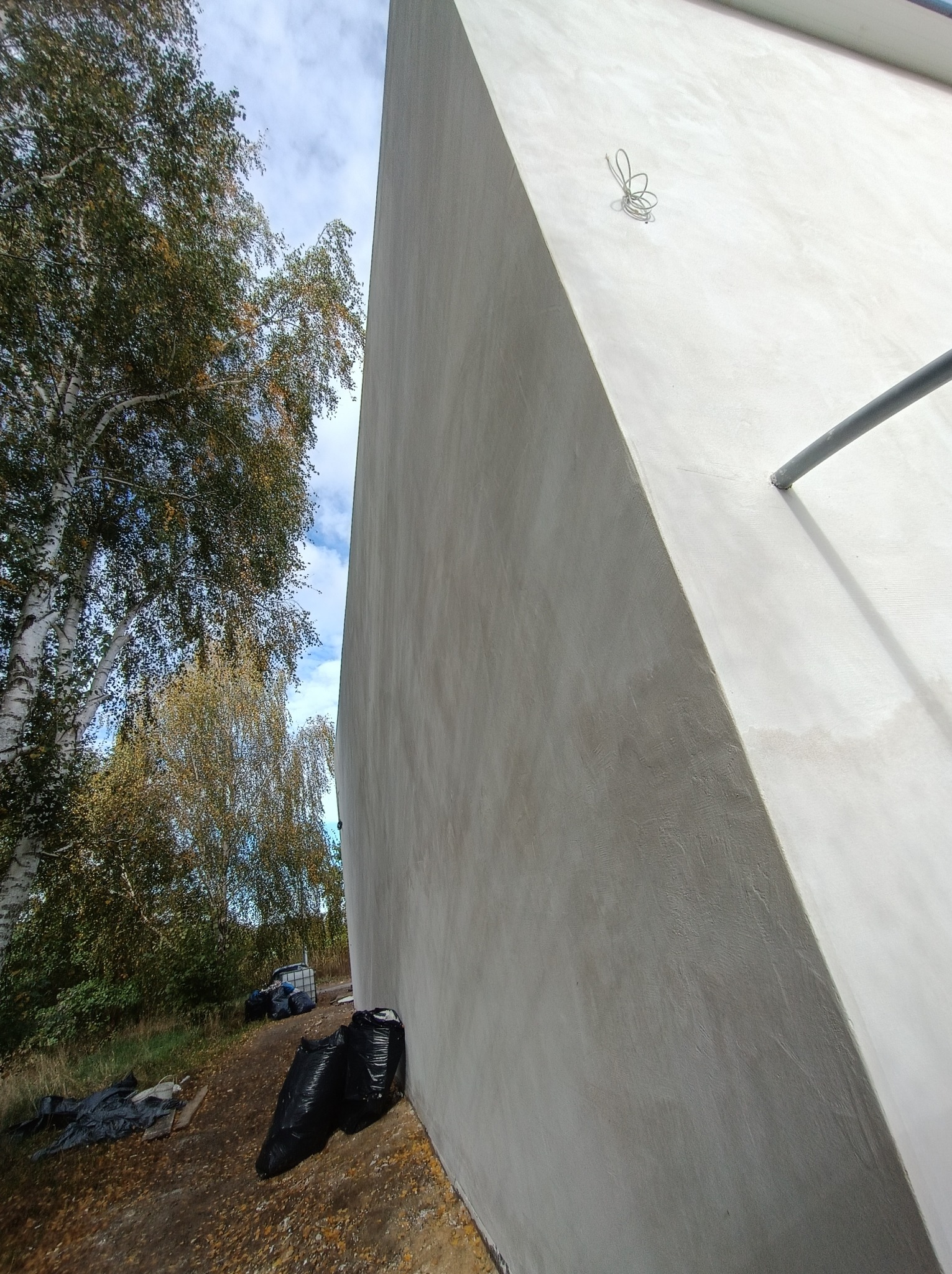 Świeżo otynkowana ściana budynku w surowym stanie, widoczne niedoskonałości faktury, obok drzewa z jesiennymi liśćmi i worki ze śmieciami budowlanymi.