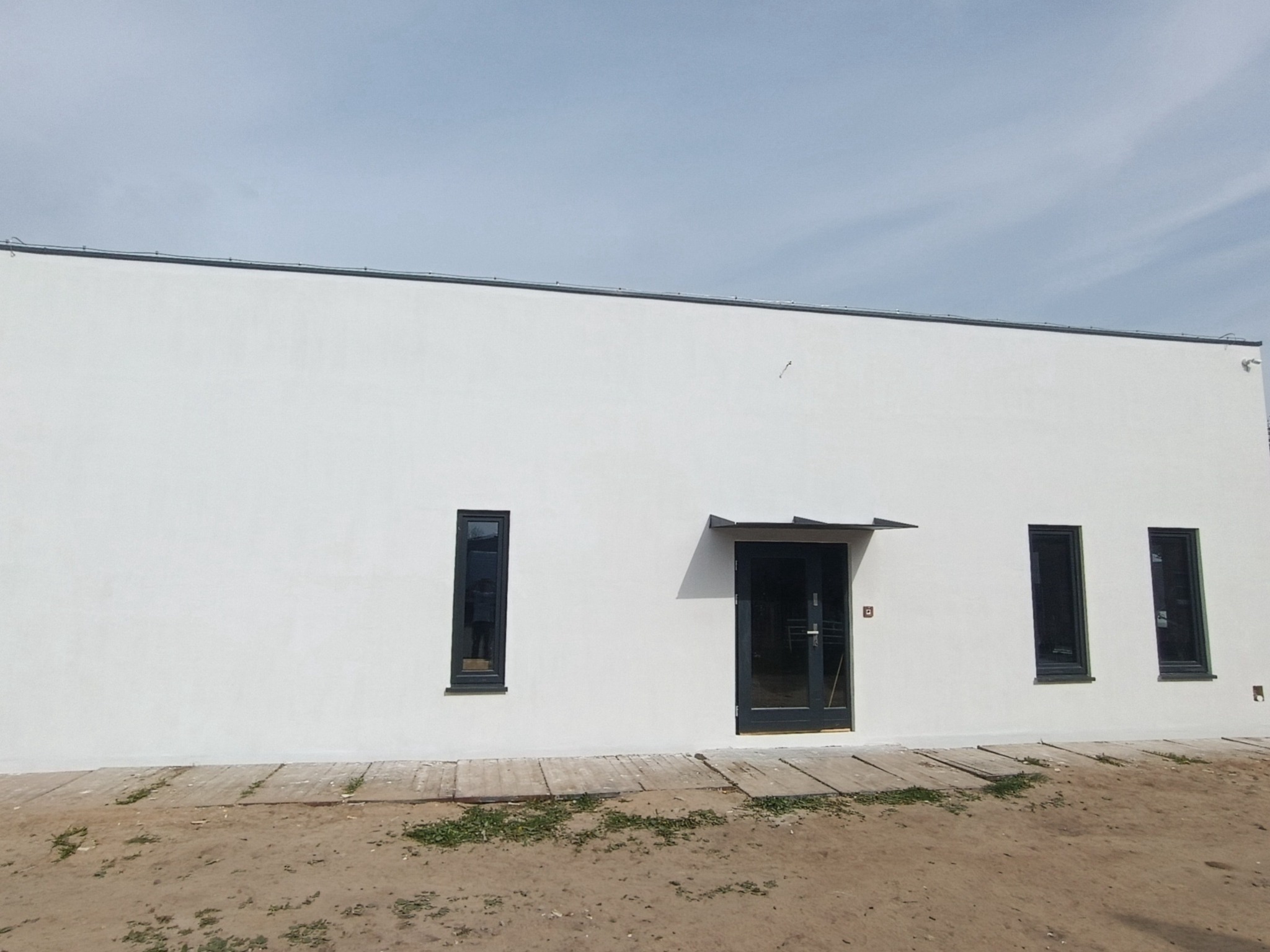 Biała elewacja budynku z ciemnymi oknami i drzwiami. Widoczne deski i piasek u podstawy, sugerujące prace wykończeniowe. Minimalistyczny design.