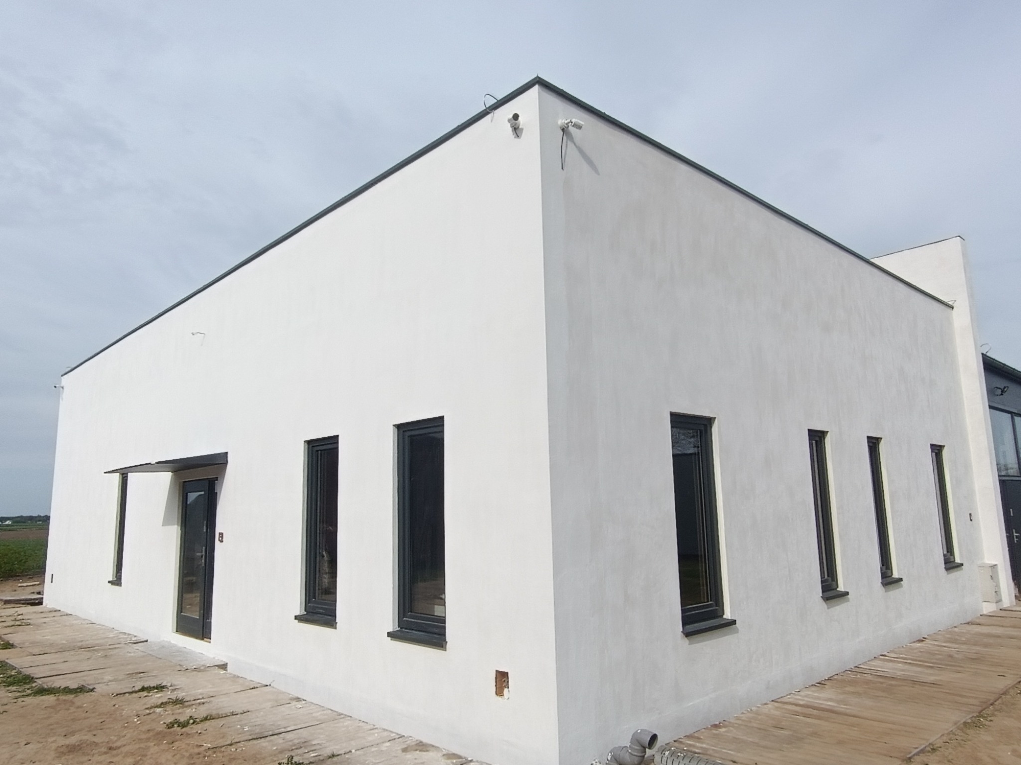 Biały budynek z ciemnymi oknami i czarnym obramowaniem dachu, widoczny z zewnątrz, z dwoma kamerami bezpieczeństwa na rogu. Minimalistyczna elewacja.