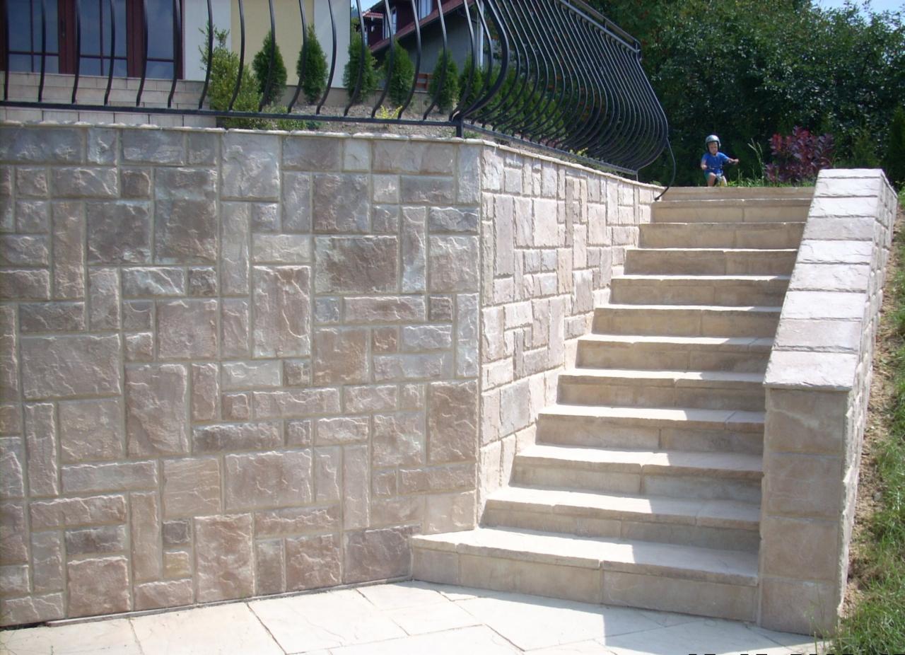 Wykończona ściana oporowa i schody zewnętrzne z betonu dekoracyjnego imitującego kamień, z czarną metalową balustradą w tle i widocznym dzieckiem w górnej części schodów.