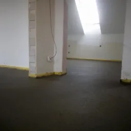 Świeżo wylana, ciemna posadzka w surowym wnętrzu z widocznymi słupami konstrukcyjnymi i żółtą taśmą na styku ściany i podłogi, oświetlona przez okno dachowe.