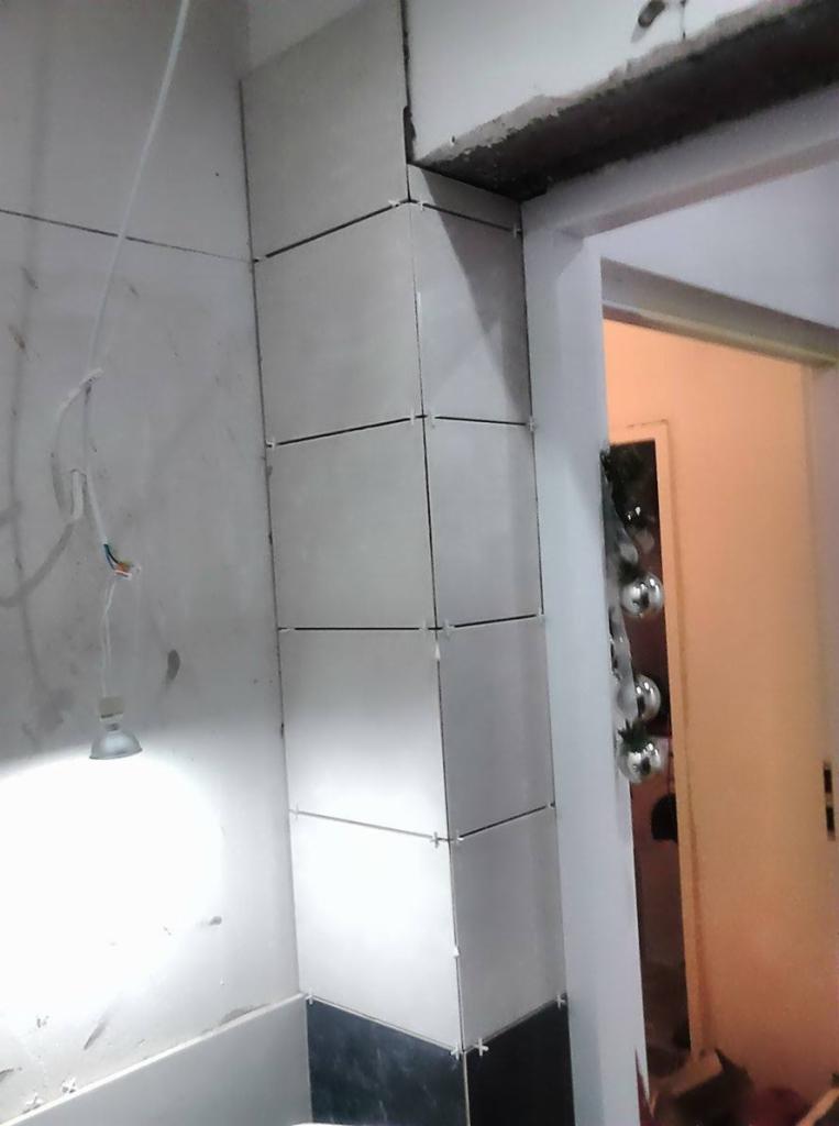 Fragment ściany z ułożonymi, jasnymi płytkami ceramicznymi. Widoczne krzyżyki dystansowe i przewody oświetleniowe. Dolna część ściany wykończona czarnymi płytkami.