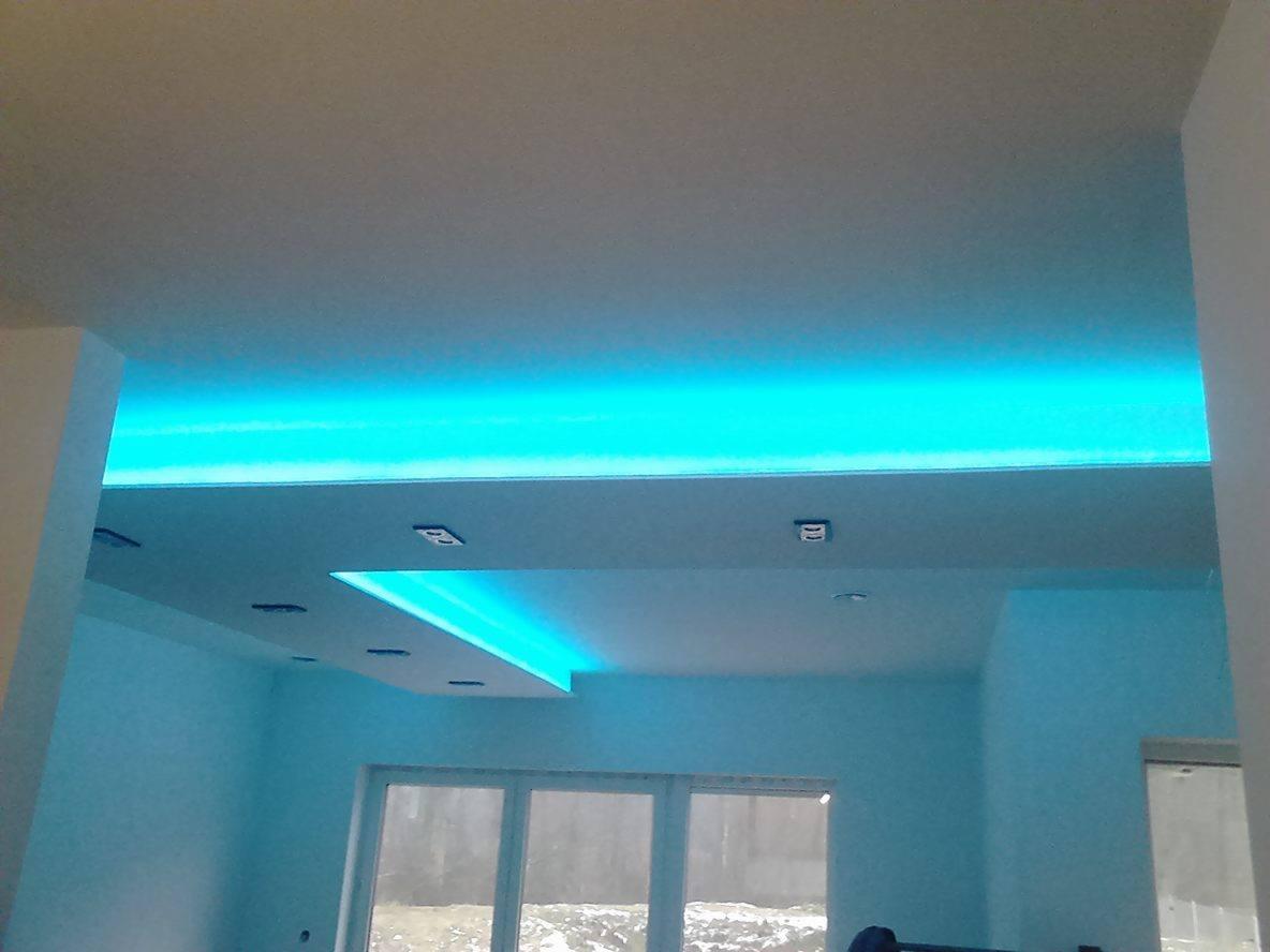 Nowoczesne oświetlenie sufitowe LED w kolorze turkusowym, podkreślające architekturę wnętrza. Widoczne okno i fragment ściany w jasnym odcieniu.