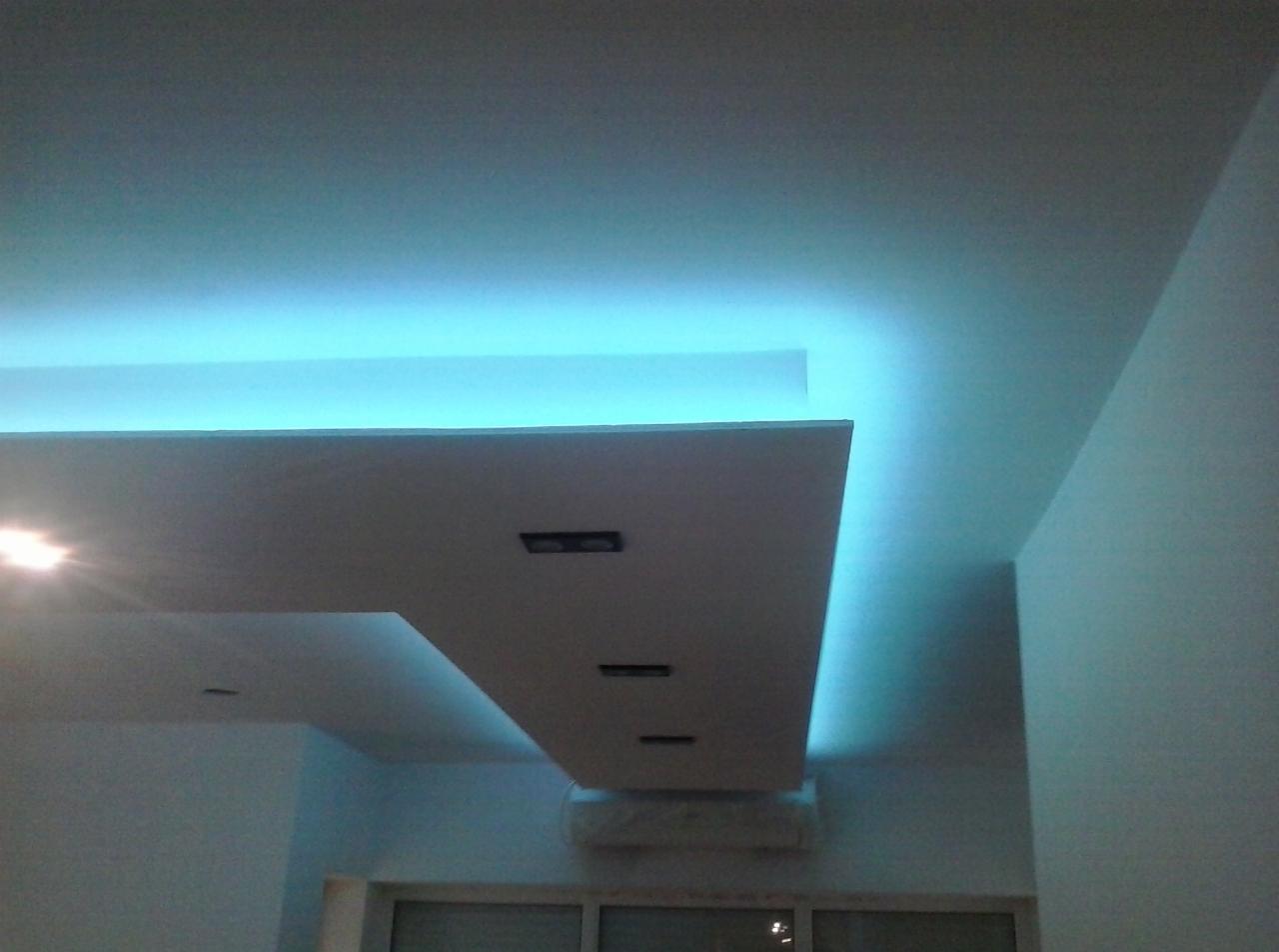 Podwieszany sufit z oświetleniem LED w kolorze chłodnego błękitu. Widoczne elementy konstrukcyjne i fragment okna w tle. Nowoczesny design wnętrza.