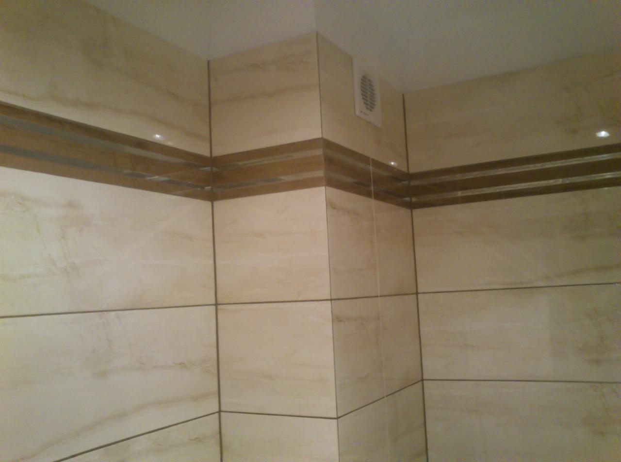Beżowe, duże płytki imitujące marmur z dekoracyjnym, brązowym pasem w wykończonej łazience. Widoczny fragment wentylacji i oświetlenie punktowe.