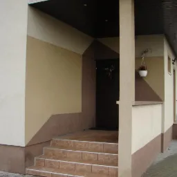 Wejście do domu z elewacją w odcieniach beżu i brązu, schody wyłożone płytkami ceramicznymi, w drzwiach wieniec.