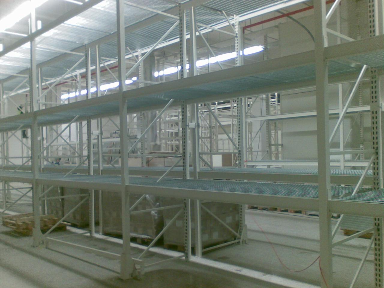 Wielopoziomowy, metalowy regał magazynowy z perforowanymi półkami, częściowo załadowany kartonami na paletach, widok z dołu, oświetlenie magazynowe.