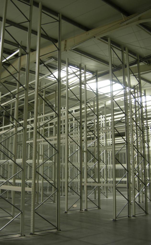 Wnętrze hali magazynowej z zamontowanymi, pustymi regałami stalowymi z kratownicami, oświetlonymi od góry przez świetliki dachowe.