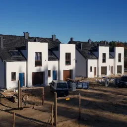 Nowoczesne, szeregowe domy z białymi elewacjami i ciemnymi dachami w trakcie budowy, widoczne samochody dostawcze i materiały budowlane na placu.