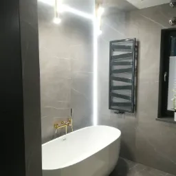 Nowoczesna łazienka z wolnostojącą białą wanną, złotą armaturą, szarą płytką imitującą marmur, podświetleniem LED i grzejnikiem drabinkowym.