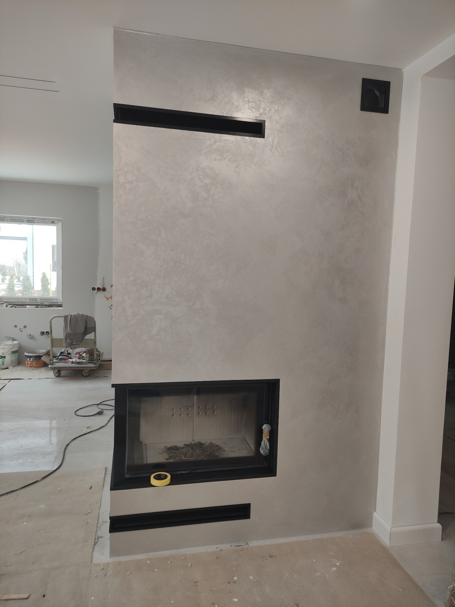 Ściana z efektem dekoracyjnym w odcieniach szarości, z wbudowanym kominkiem z czarną obudową i elementami wentylacyjnymi, widoczne prace wykończeniowe w tle.