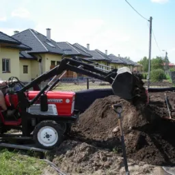 Czerwony traktor z łyżką nabiera ziemię na budowie, widoczny operator w okularach przeciwsłonecznych, w tle szereg domów jednorodzinnych.