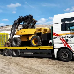 Żółta koparka Mecalac przewożona na niskopodwoziowej lawecie z żółtymi burtami, ciągniętej przez białą ciężarówkę z napisem 'AUTO-HOLOWANIE' na tle błękitnego nieba.