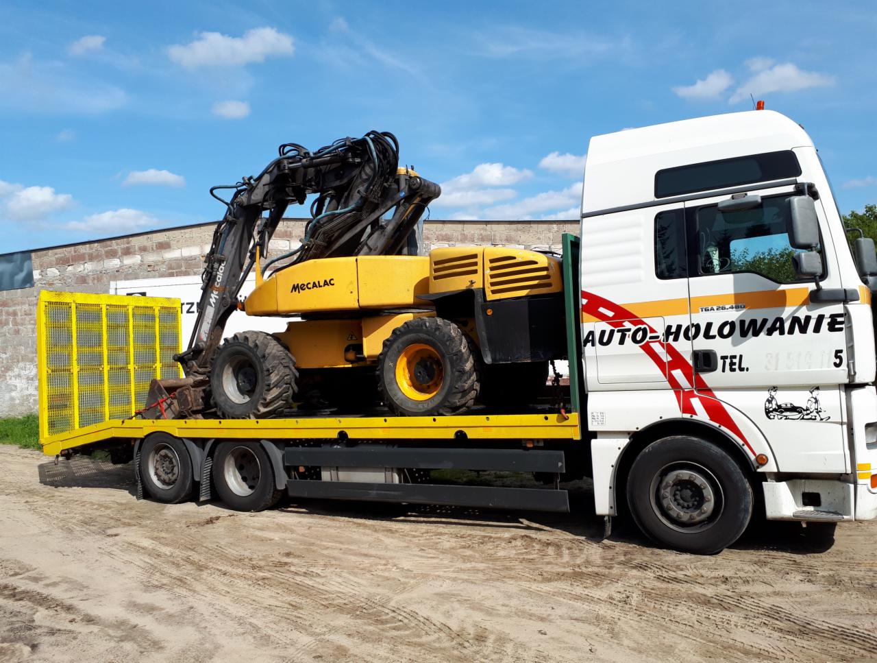 Żółta koparka Mecalac przewożona na niskopodwoziowej lawecie z żółtymi burtami, ciągniętej przez białą ciężarówkę z napisem 'AUTO-HOLOWANIE' na tle błękitnego nieba.