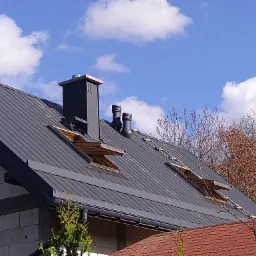 Szary dach z blachy trapezowej z trzema oknami dachowymi, kominem i dwiema rurami wentylacyjnymi na tle błękitnego nieba z chmurami.