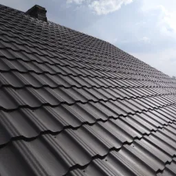 Nowe, ciemnoszare pokrycie dachowe z blachodachówki modułowej, widok z dołu pod kątem, komin w tle, błękitne niebo z chmurami.